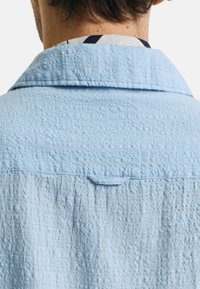 Camicia tessuta azzurro chiaro con colletto ripiegato, caratterizzata da un motivo a trapunta discreto e un asola sul retro per appenderla.
