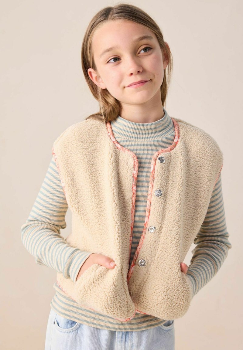 Gilet en velours crème avec des détails en pêche, comportant des boutons-pression et des poches avant, porté sur un col roulé rayé bleu clair et blanc.