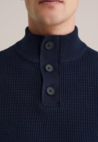 Mörkblå stickad tröja med ett texturerat mönster, hög krage och tre svarta knappdetaljer vid halslinningen, vilket visar en närbild.