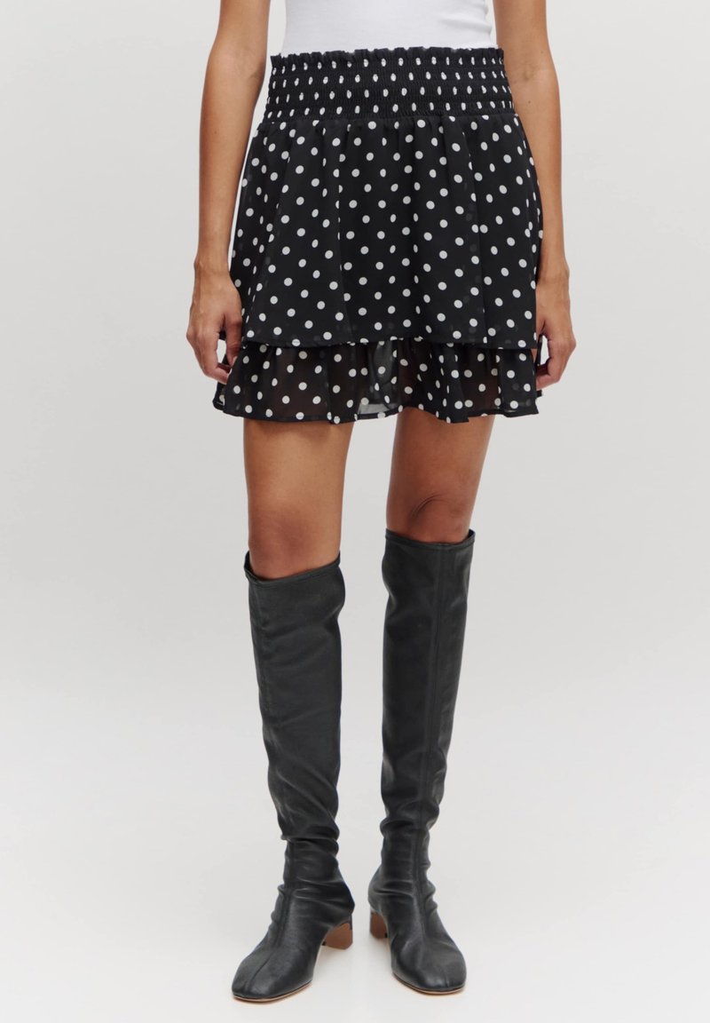 Zwarte rok met stippenpatroon en gelaagd ontwerp, met smocktaille, gemaakt van lichte stof. Gecombineerd met zwarte laarzen tot de knie met hak.