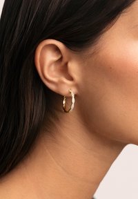 Pendiente de aro dorado con un diseño retorcido, adornado con pequeños cristales transparentes a lo largo de un lado, mostrado en una oreja.