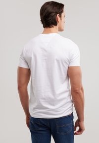 Uomo che indossa una maglietta bianca a maniche corte e jeans blu, in piedi con le spalle rivolte verso la fotocamera contro uno sfondo grigio chiaro.