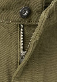 Tela de mezclilla verde oliva con cierre de cremallera metálica y botón. La textura es suave con detalles de costura visibles y un aspecto resistente.