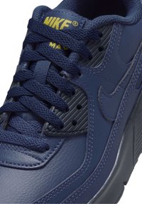 Gros plan d'une basket Nike Air Max bleu marine avec un logo jaune, cuir texturé et lacets épais bleu marine noués sur le dessus.