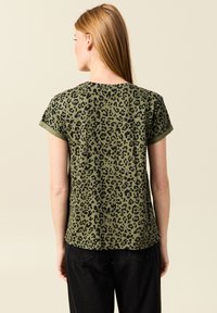 Camiseta de manga corta en verde oliva con un patrón de estampado de leopardo negro. Presenta un corte relajado y mangas con puños para un detalle adicional.