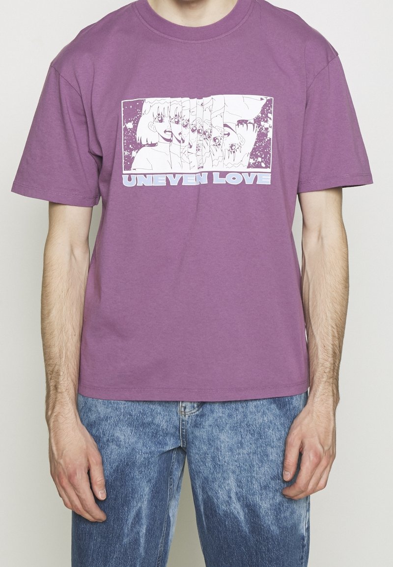 Personne portant un t-shirt violet avec un graphique blanc d’un visage de style anime fragmenté et le texte "UNEVEEN LOVE" en lettres capitales.