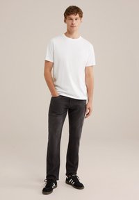 Baltas marškinėliai su apvaliu kaklu, juodi slim-fit džinsai, juodi zomšiniai bateliai su baltais dryžiais. Neutrali fono spalva, modelis stovi su viena ranka kišenėje.