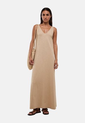 Femme portant une longue robe beige sans manches, portant un sac à bandoulière tressé et des sandales marron foncé, debout devant un fond clair.