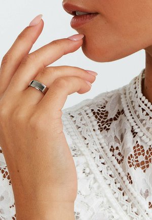Sølv ring med en poleret overflade, båret på en hånd, der hviler mod læberne, pyntet med en hvid blondebluse med detaljerede mønstre.