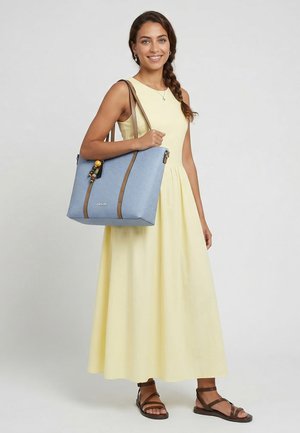 Femme en robe jaune sans manches tenant un grand sac fourre-tout bleu avec des anses marron, portant des sandales marron et souriant.