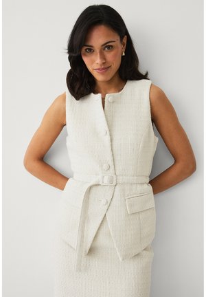 Kobieta o ciemnych włosach ubrana w białą, bezrękawnikową, teksturowaną sukienkę z guzikami i paskiem w talii, pozująca na tle jednolitej białej ściany.