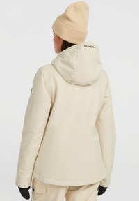 Beige pufferjack met capuchon, met een zachte textuur en een logo op de achterkant. Het model draagt een lichtgekleurde gebreide muts en handschoenen.
