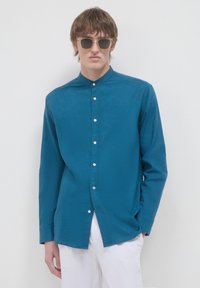 Camicia teal con colletto alla cinese, maniche lunghe e bottoni bianchi; modello abbinato a pantaloni chiari e occhiali da sole.