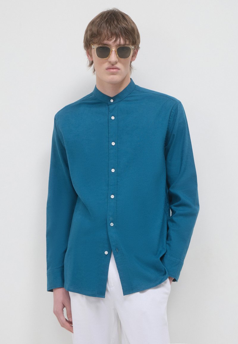 Camicia teal con colletto alla cinese, maniche lunghe e bottoni bianchi; modello abbinato a pantaloni chiari e occhiali da sole.