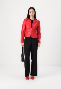 Rote Lederjacke mit Reißverschluss, kombiniert mit schwarzen weiten Hosen und roten Absatzschuhen. Hält eine kleine schwarze Tasche. Einfarbiger Hintergrund.