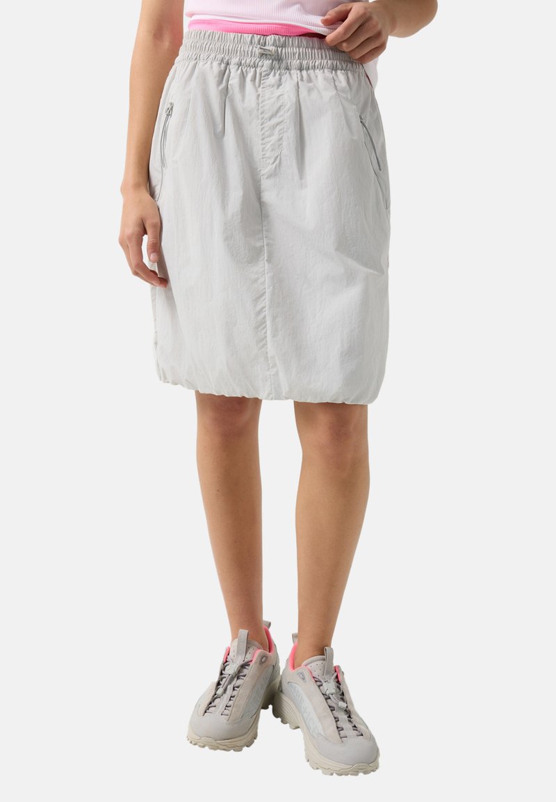 Mujer con falda gris claro hasta la rodilla con cinturilla elástica y bolsillos con cremallera, combinada con zapatillas deportivas de color gris claro y rosa.