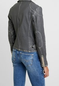 Kvinde iført en tætsiddende grå læderjakke med lynlåsmansjetter og blå jeans, vist fra ryggen mod en ensfarvet baggrund.