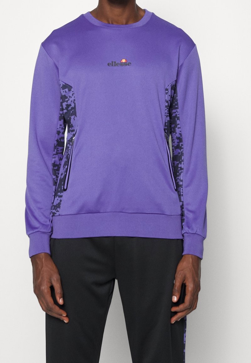 Homme portant un sweat-shirt à manches longues violet avec des panneaux en camouflage numérique et des poches à fermeture éclair noires, associé à un pantalon noir.