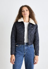 Chaqueta negra acolchada con un cuello de piel sintética crema, cierre de cremallera y dos bolsillos delanteros, combinada con unos vaqueros azules de cintura alta.