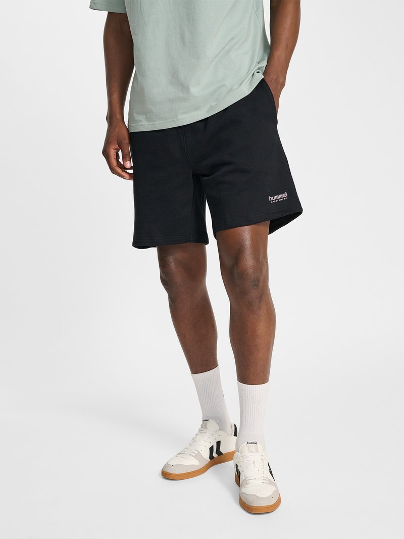 Mann trägt hellgrünes T-Shirt, schwarze Hummel-Shorts, weiße Knöchelsocken und weiße Sneaker mit schwarzen und Gummisohlen, steht mit einer Hand in der Tasche.