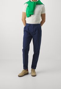 Man in beige sneakers, navy blauwe broek, wit polo shirt en een groene trui over zijn schouders, tegen een plain witte achtergrond.