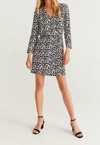 Schwarz-weißes Kleid mit Wickeldesign, langen Ärmeln und Blattmuster. Kombiniert mit schwarzen Riemchen-Absätzen. Glatte Stoffoberfläche.
