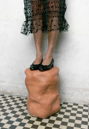 Ballerine in pelle nera con dettaglio a fiocco, posizionate su una base in scultura terracotta testurizzata, su un pavimento a scacchi bianco e nero.