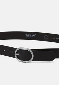 Ceinture en cuir noir avec une boucle ovale en argent. Présente un design minimaliste, une surface texturée et le logo embossé "Levi's" près de la boucle.