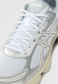 Närbild av en vit och silver ASICS löparsko med vita snören och en beige sula på en vit bakgrund.