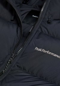 Sort pufferjakke med en glat, mat overflade. Har en frontlynlås og broderet "Peak Performance" logo på brystet.
