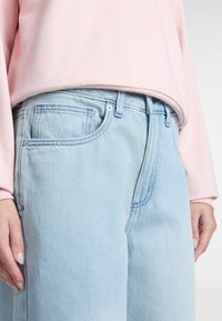 Jeans in denim azzurro chiaro con una vestibilità comoda, dotati di cinque tasche e cuciture sottili. Indossati con un morbido top a maniche lunghe rosa.