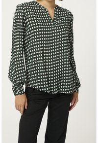 Zielona bluzka w wzory z długimi rękawami, z dekoltem w serek i diamentowymi kształtami w białym i ciemnozielonym kolorze, noszona z czarnymi spodniami.