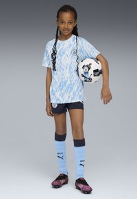 Gaiši zils un balts rakstains krekls ar Mančestras City logo, tumši zili šorti, zili un melni zeķes, melni un rozā futbola apavi, tur rokās futbola bumbu.