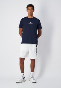 Camiseta de algodón en azul marino oscuro con un pequeño logo de Champion, combinada con pantalones cortos de felpa blancos con letra en los laterales. Unos zapatos deportivos y calcetines completan el look.