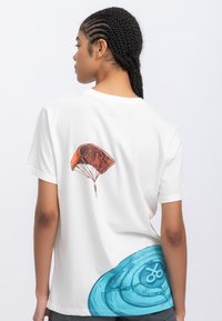 T-shirt bianca in cotone con grafica di un paracadute rosso e un motivo a spirale blu sul retro, con maniche corte e scollatura a girocollo.
