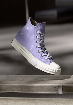 Lawendowy sneaker na wysokiej cholewce z białymi sznurowadłami i kwiatowymi wzorami, unoszący się nad betonową powierzchnią z miękkimi cieniami.