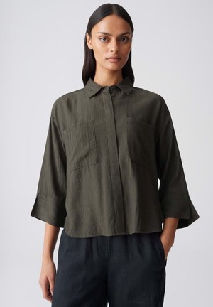 Femme aux longs cheveux foncés portant une chemise texturée vert olive à boutons avec des manches larges et un pantalon noir, debout sur un fond neutre.