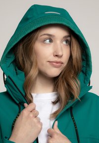 Tealfarbene Kapuzenjacke mit glatter Oberfläche, ausgestattet mit einem Reißverschluss, verstellbaren Kordeln und einem kontrastierenden Innenfutter. Das Modell trägt ein weißes Oberteil.