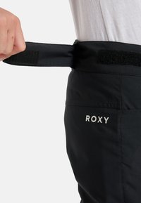 Czarne spodnie z regulowanym pasem na rzep, z białym haftem logo "ROXY" z boku. Gładka struktura materiału.