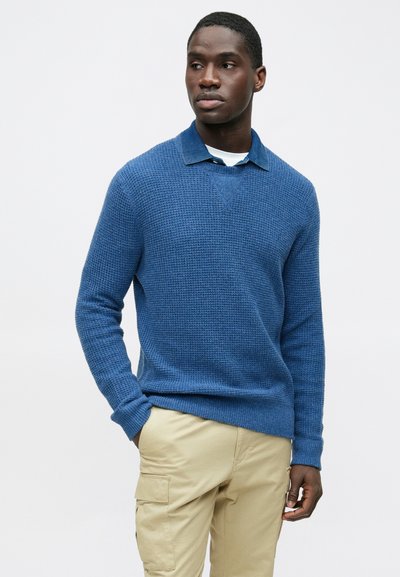 Mėlynas megztas megztinis su tekstūruotu audiniu, su apykakle ant baltos marškinių, dėvimas su smėlio spalvos kargo kelnėmis.