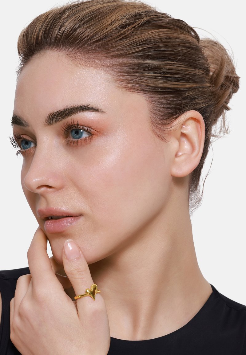 Anello in tonalità oro a forma di cuore, indossato sull'indice. Il design presenta curve morbide e una finitura lucida su uno sfondo neutro.