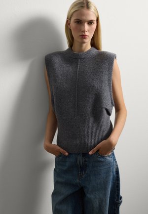 Femme blonde portant un pull gris sans manches avec épaulettes et un jean bleu ample, se tenant contre un mur blanc les mains dans les poches.