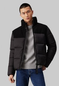 Giacca a vento nera con colletto alto, caratterizzata da un pannello nero dalla texture morbida nella parte superiore, chiusura con zip e tasche laterali. Indossata su un maglione grigio.