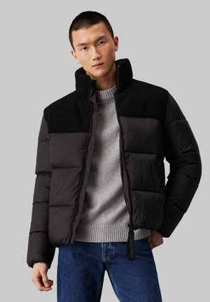 Chaqueta de plumas negra con collar alto, que presenta un panel negro de textura suave en la parte superior, cierre con cremallera y bolsillos laterales. Llevada sobre un suéter gris.