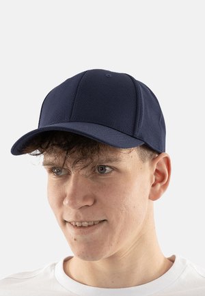 Giovane uomo con capelli ricci che indossa un berretto da baseball blu scuro e una camicia bianca, guardando leggermente verso destra con un sorriso.