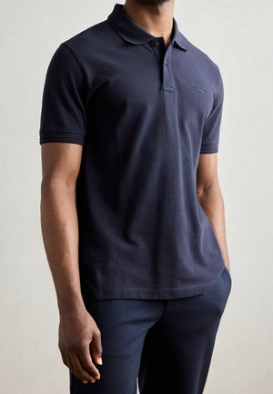 Polo bleu marine en tissu de coton avec un col classique, deux boutons et un petit logo sur la poitrine ; porté avec un pantalon assorti.