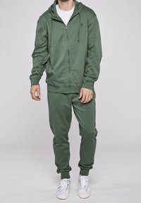 Uomo in felpa verde oliva con zip e pantaloni jogger abbinati, indossando sneakers bianche, su sfondo neutro.