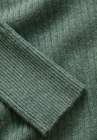 Tissu vert côtelé avec une surface texturée, doté d'un revers légèrement allongé. Le matériau semble doux et confortable.