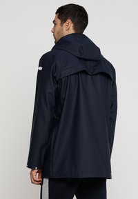 Navy waterproof jacket with a hood, smooth texture, adjustable cuffs, and logo on the shoulder. Features side pockets and a relaxed fit.