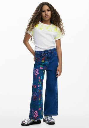 Valge T-särk neoonselt kollaste mustriliste varrukatega, kombineeritud siniste lehvikpükstega, millel on värvilised graafilised kujundused. Välimust täiustavad hõbedased sandaalid.